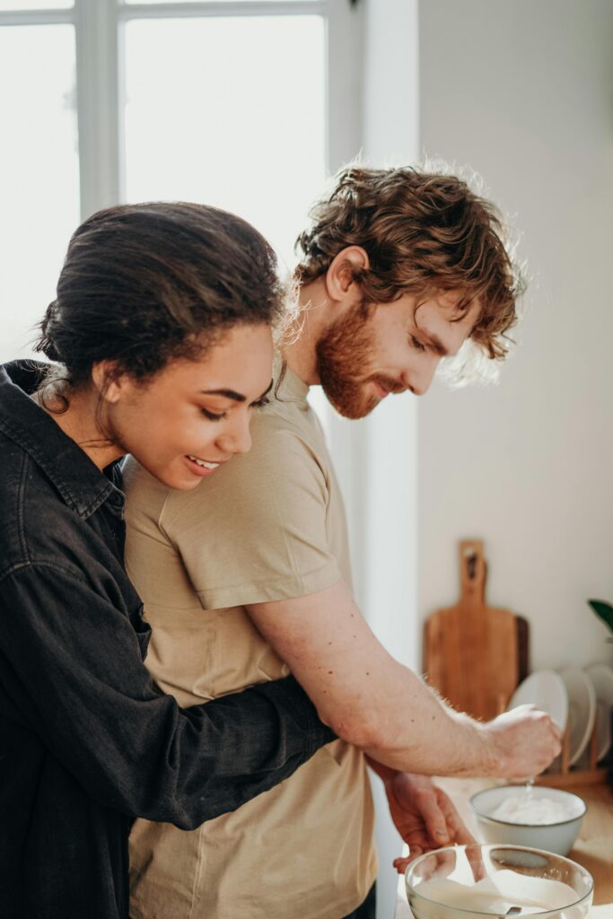 Wife smiling and hugging her husband from behind as he cooks, while both enjoying an experiential intimate moment